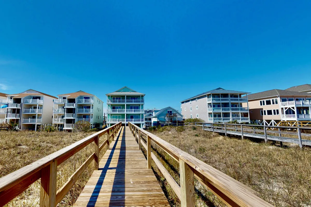 A cheerful exterior shot featuring a classic wooden boardwalk with beach houses in the background and a bright, blue sky overhead. The scene suggests a lively, welcoming coastal community and easy access to the beach for outings with pets.