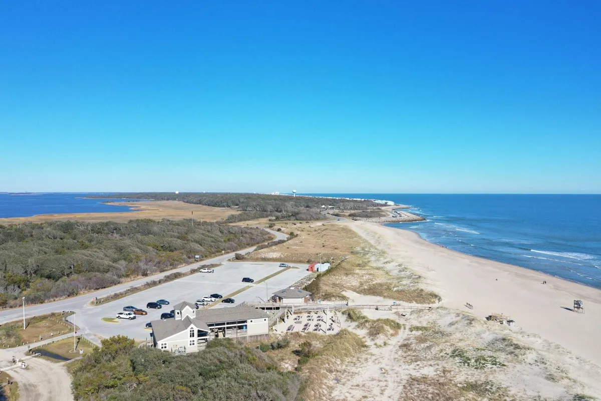 This image captures the exterior of a coastal building with a visible parking area in the foreground, lush vegetation, and the beach and ocean just beyond. The relaxed scene suggests convenience for arriving guests, with easy access to both parking and the shoreline.