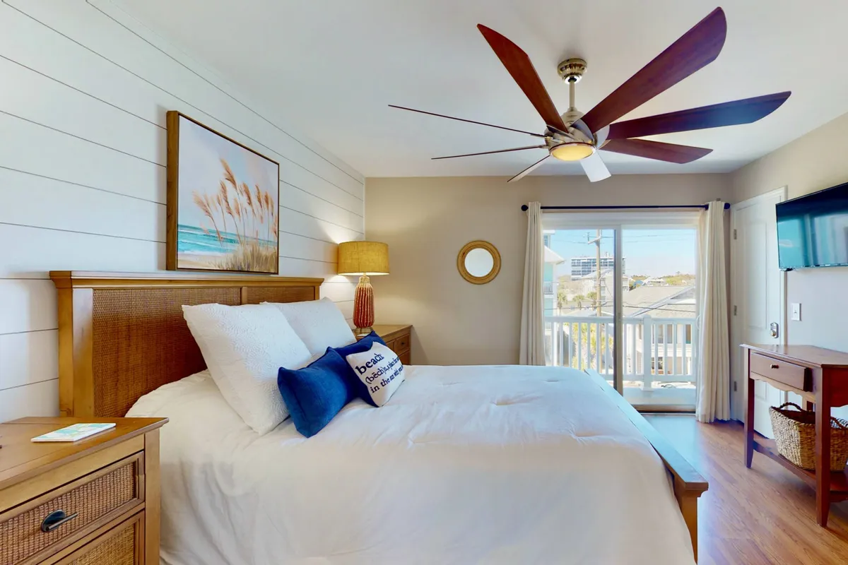 A spacious bedroom featuring a queen bed with elegant linens, ceiling fan, wall art, and large glass doors that open to a balcony or offer ocean views. Natural light pours in, and the room feels airy and peaceful—perfect for either couples or families seeking rejuvenation.