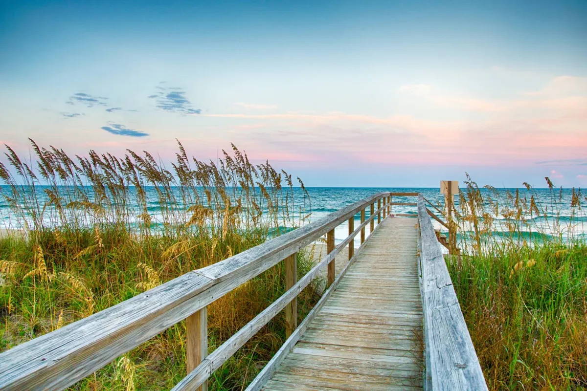 A serene scene of a wooden boardwalk stretching across dune grass toward the ocean, with gentle waves and a colorful sunset sky in the background. This peaceful view highlights the natural beauty of the coastal environment and the inviting approach to the beach.