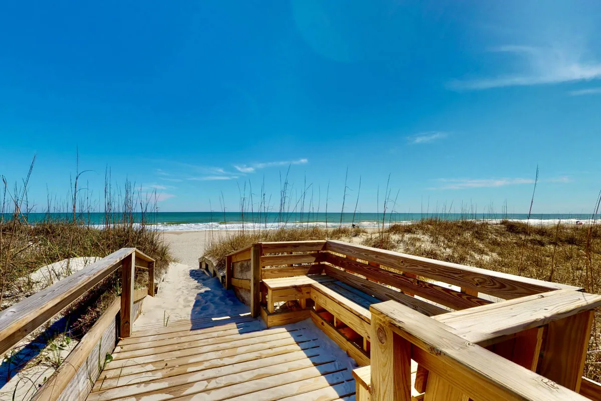 A wooden walkway leads directly to a wide, sandy beach and the sparkling ocean beyond. A cozy seating area offers a family-friendly spot for relaxing, watching the sunrise, or keeping an eye on kids building sandcastles just steps away from the waves.