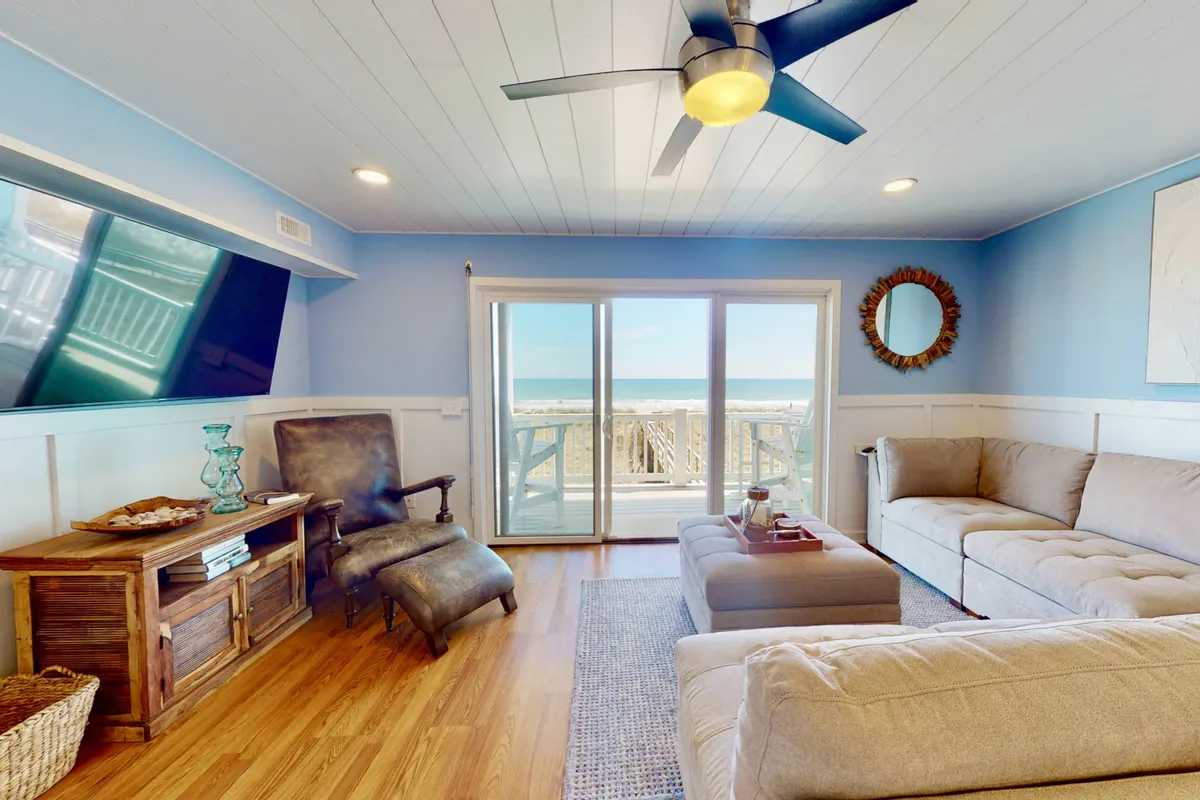 A spacious, sunlit living area with a comfortable sectional sofa facing large windows that frame a stunning ocean view. The room features a wooden coffee table, a flat-screen TV, and a decorative mirror, with sliding doors that open directly onto the private deck. The ample seating and open sightlines create a casual, welcoming atmosphere designed for both relaxation and group gatherings.