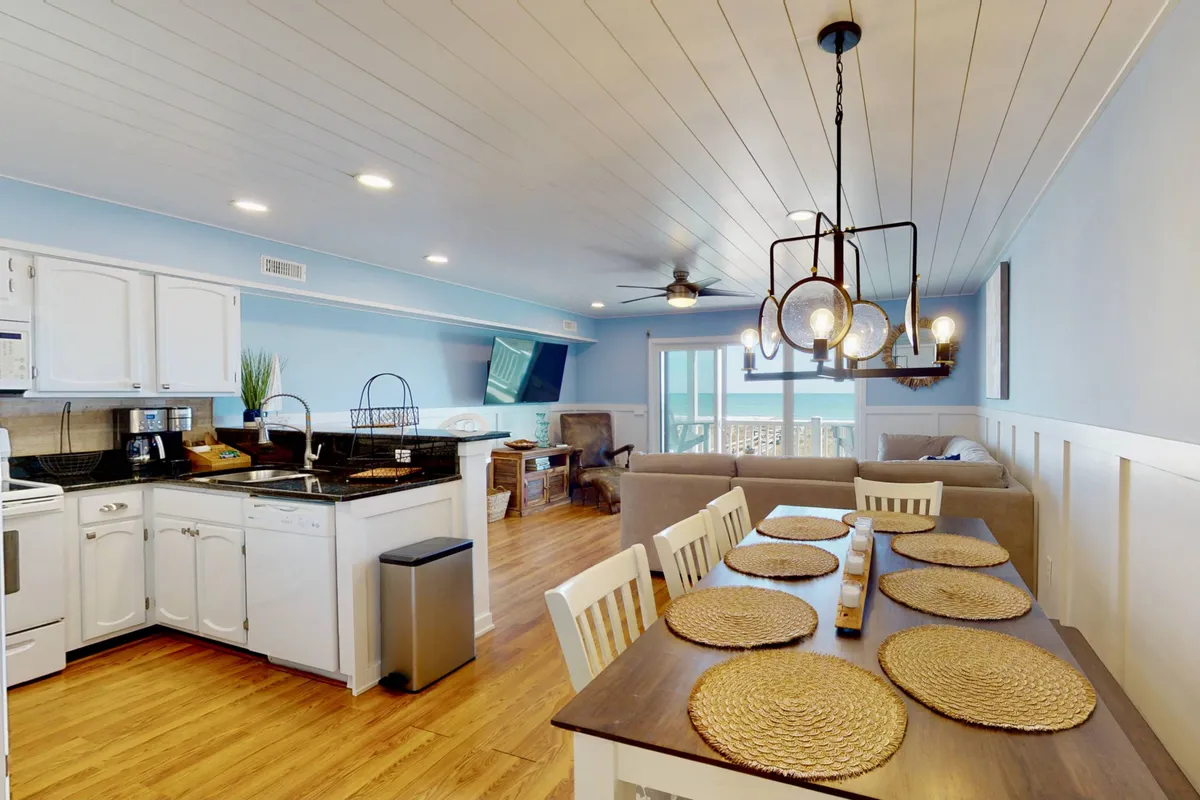 A bright, inviting open-concept space that seamlessly connects the dining area, kitchen counter, and adjacent living area seating. The modern light fixtures and carefully placed décor enhance the welcoming mood, while the proximity to the deck allows sunlight to flood the room, hinting at indoor-outdoor living.