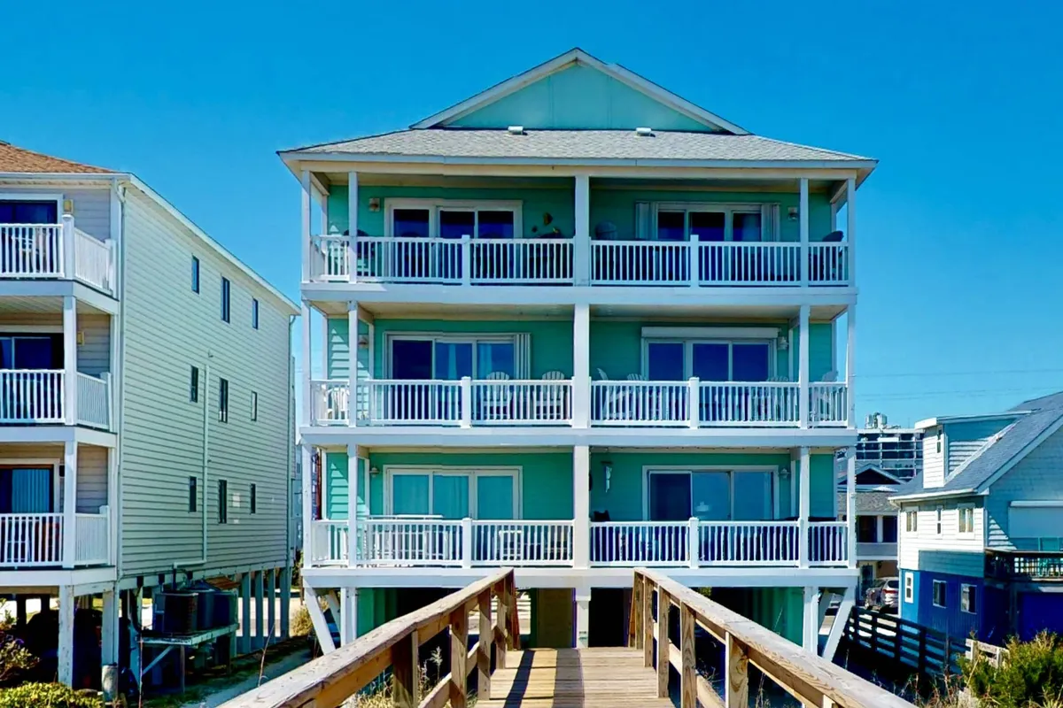 The cheerful turquoise exterior of the condo, featuring spacious balconies and a wooden walkway leading to the entrance. The image radiates a welcoming, relaxed mood and hints at easy access to the beach—key for morning strolls with your dog.