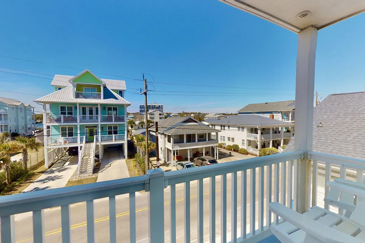 A vibrant view from the balcony features colorful beach houses, palm trees, and a clear blue sky. The scene captures the inviting atmosphere of the neighborhood and the sense of being part of a lively coastal community.