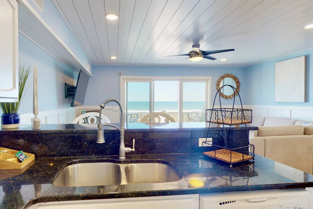A spacious, sunlit living area seamlessly connected to the kitchen, with large windows offering an ocean view. The living room features comfortable seating ideal for relaxing with your dog at your feet. The space is casual and serene, with practical flooring visible and an inviting atmosphere for both pets and people.