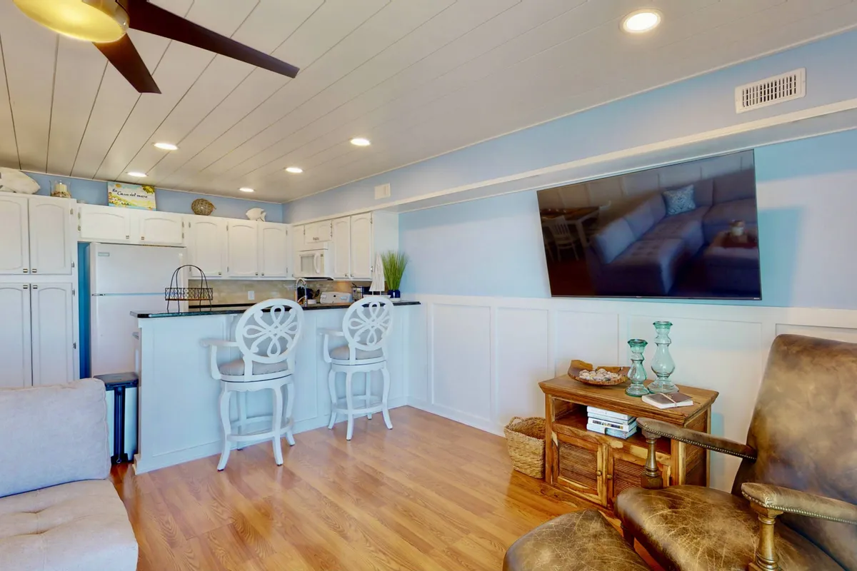 A cozy, inviting kitchen and living area with a kitchen island, bar stools, television, and comfortable armchair. The light, practical flooring is visible, and the layout is ideal for hosting, relaxing, or letting a pet lounge nearby.