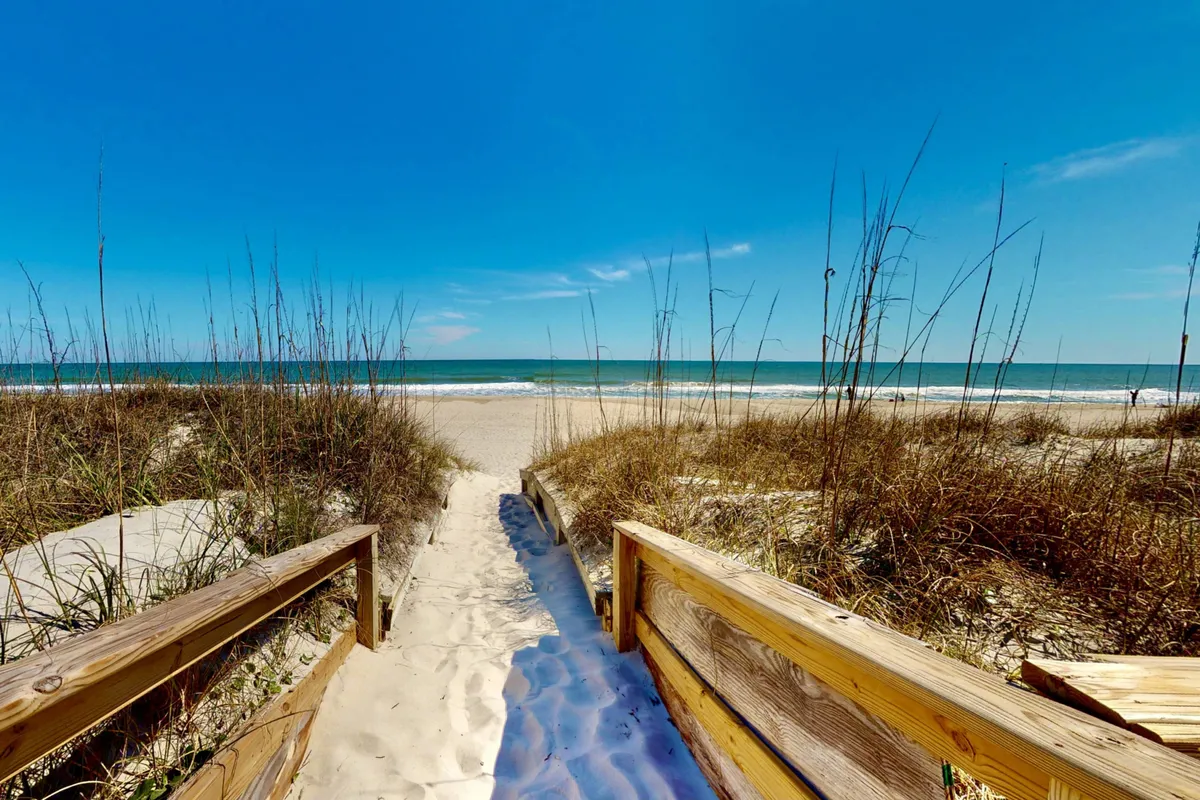 A wooden walkway leads over sandy dunes directly to the ocean, with a brilliant blue sky overhead. The scene is bright and open, evoking the feel of easy access to the beach and a tranquil work-adjacent escape.