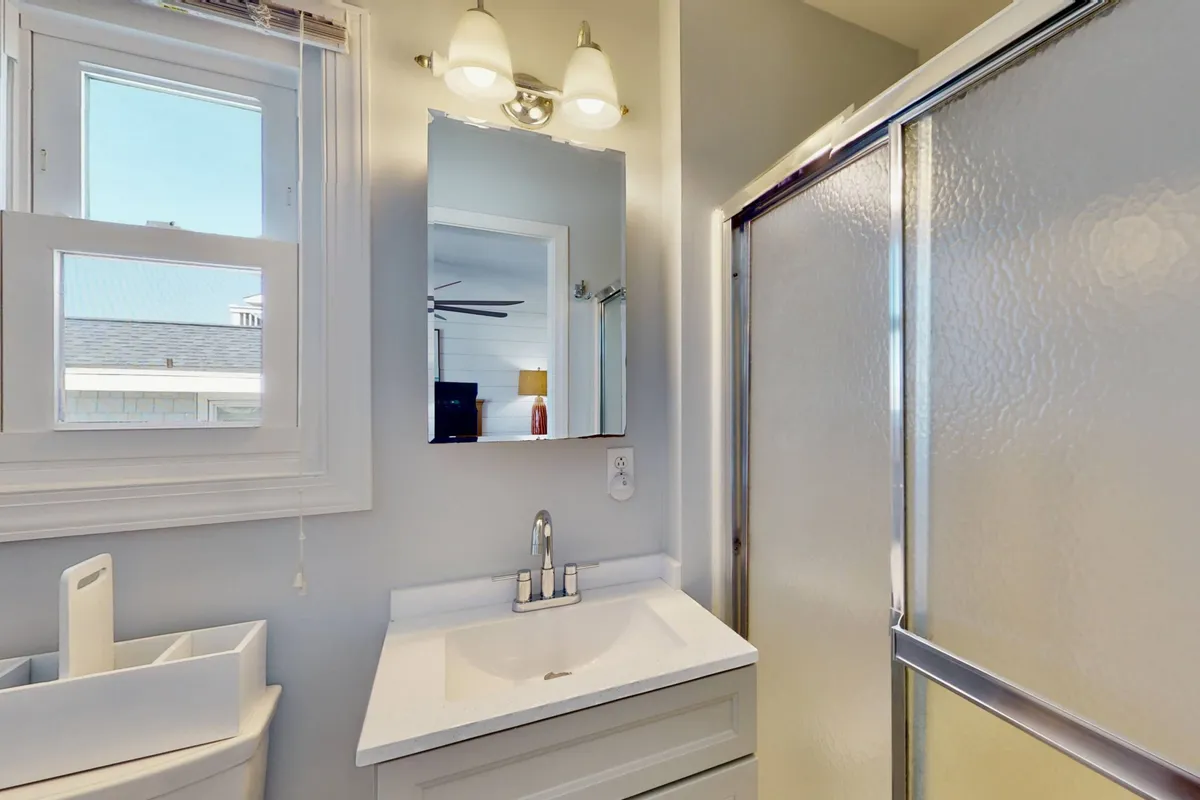 A bright, inviting bathroom with a modern sink, large mirror, walk-in shower, and a window that lets in natural light. Fluffy towels are neatly arranged, creating a spa-like environment for refreshing after a day at the beach.