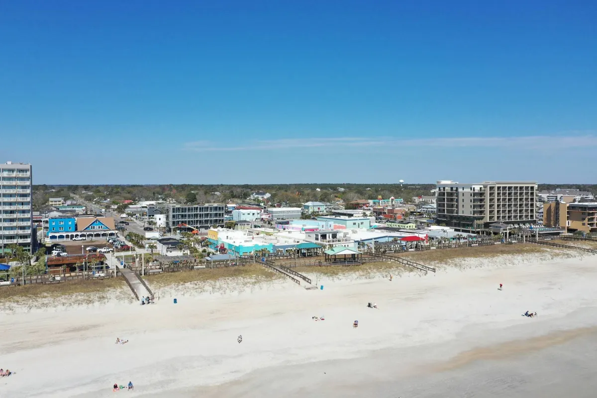 A lively view of the beachfront lined with colorful buildings and a bustling boardwalk. People are enjoying the sunny beach, swimming, sunbathing, and strolling, with shops and restaurants visible nearby, all set against the backdrop of the ocean.