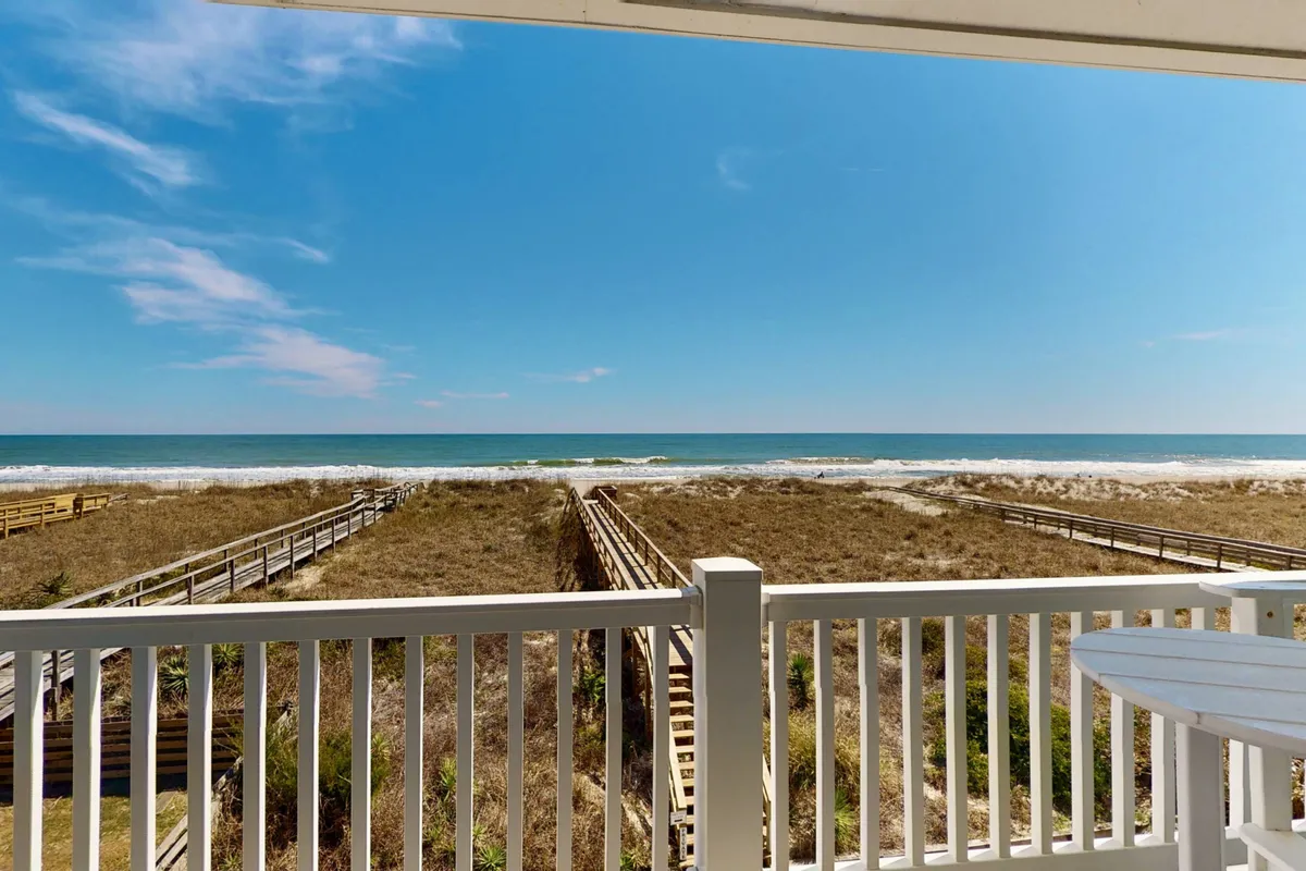 A serene view featuring the ocean, boardwalks leading across dune grass to the beach, and a wide expanse of water. The scene is peaceful yet inviting, suggesting early morning or evening relaxation on the balcony with the beach just steps away.