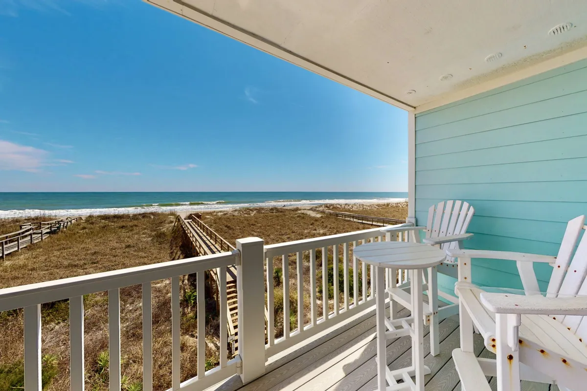 A spacious wooden deck overlooking the sparkling Atlantic Ocean, featuring two inviting white chairs perfectly positioned for relaxing and taking in sunrise or sunset views. The tranquil setting suggests a peaceful retreat, with easy access to the beach just steps away.