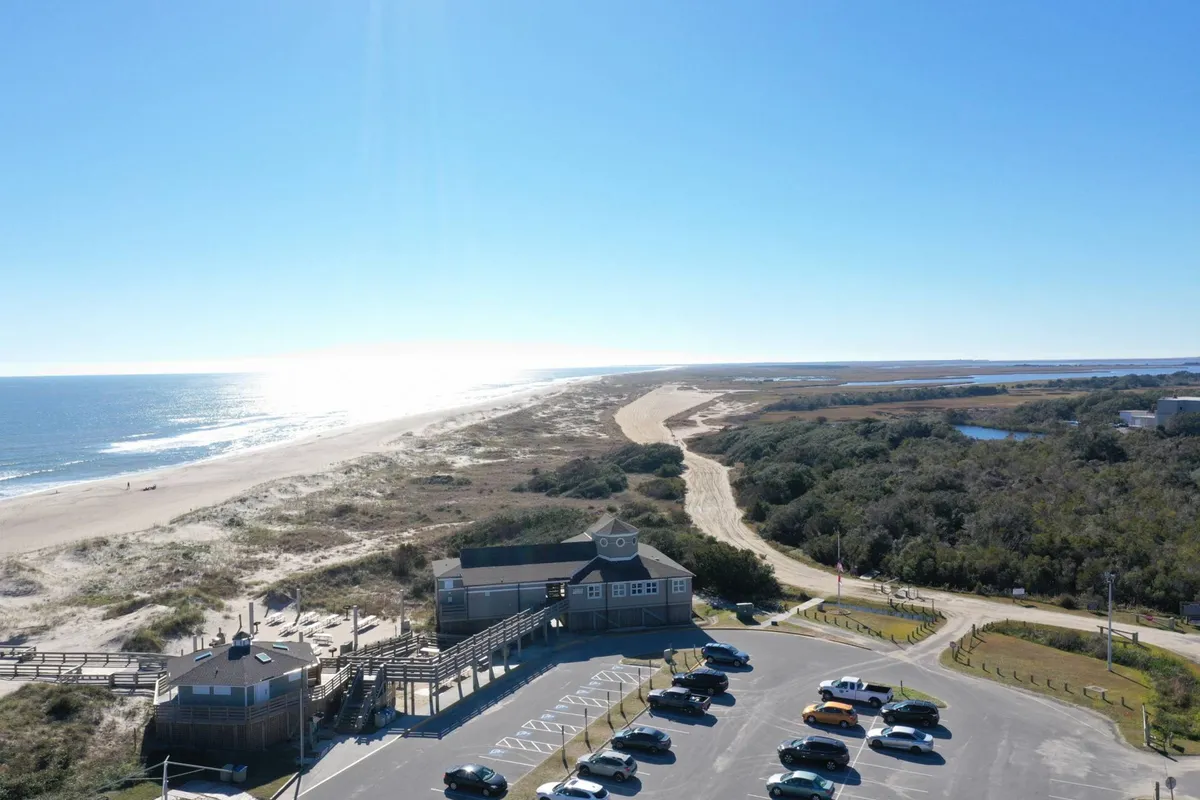 An exterior view featuring a parking lot adjacent to a beach, a pathway leading up to the building, and ocean waves in the background. The approachable entrance and visible storage possibilities connect to practical vacation needs.