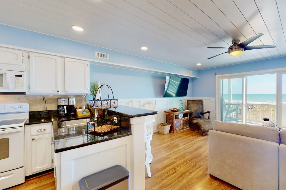 A cozy, modern kitchen with a central island provides ample space for cooking and entertaining. The kitchen opens up to a comfortable seating area and television, with expansive windows offering unobstructed views of the ocean and letting in abundant natural light.